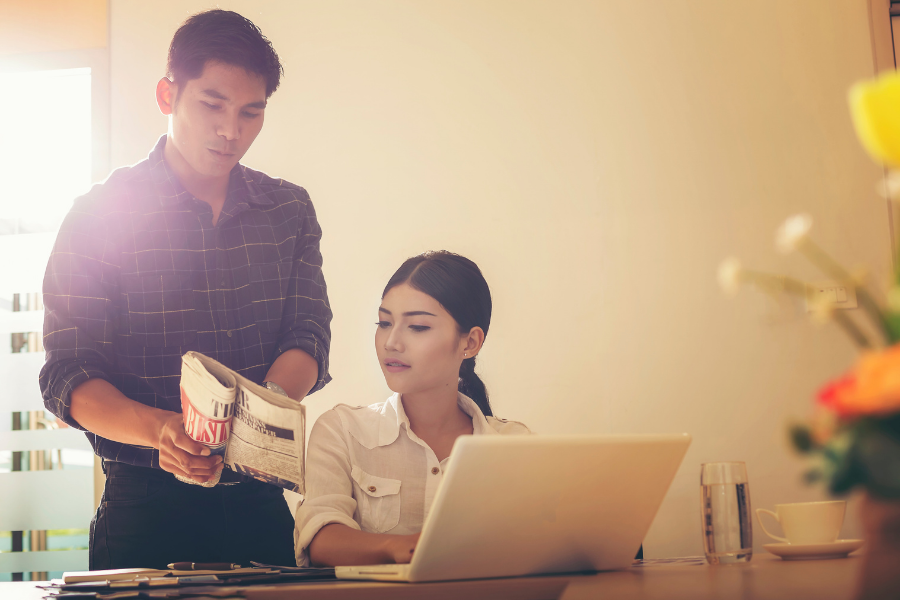 Two people reading a newspaper and looking at a laptop