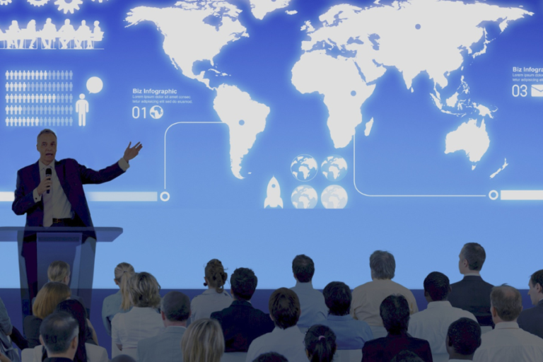 A man stands at a podium in front of an audience pointing towards a map of the world.