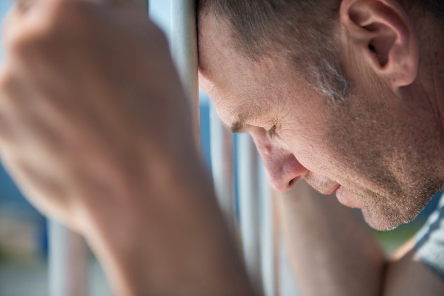 A man gripping the bars of a jail cell and resting his forehead against the bars.